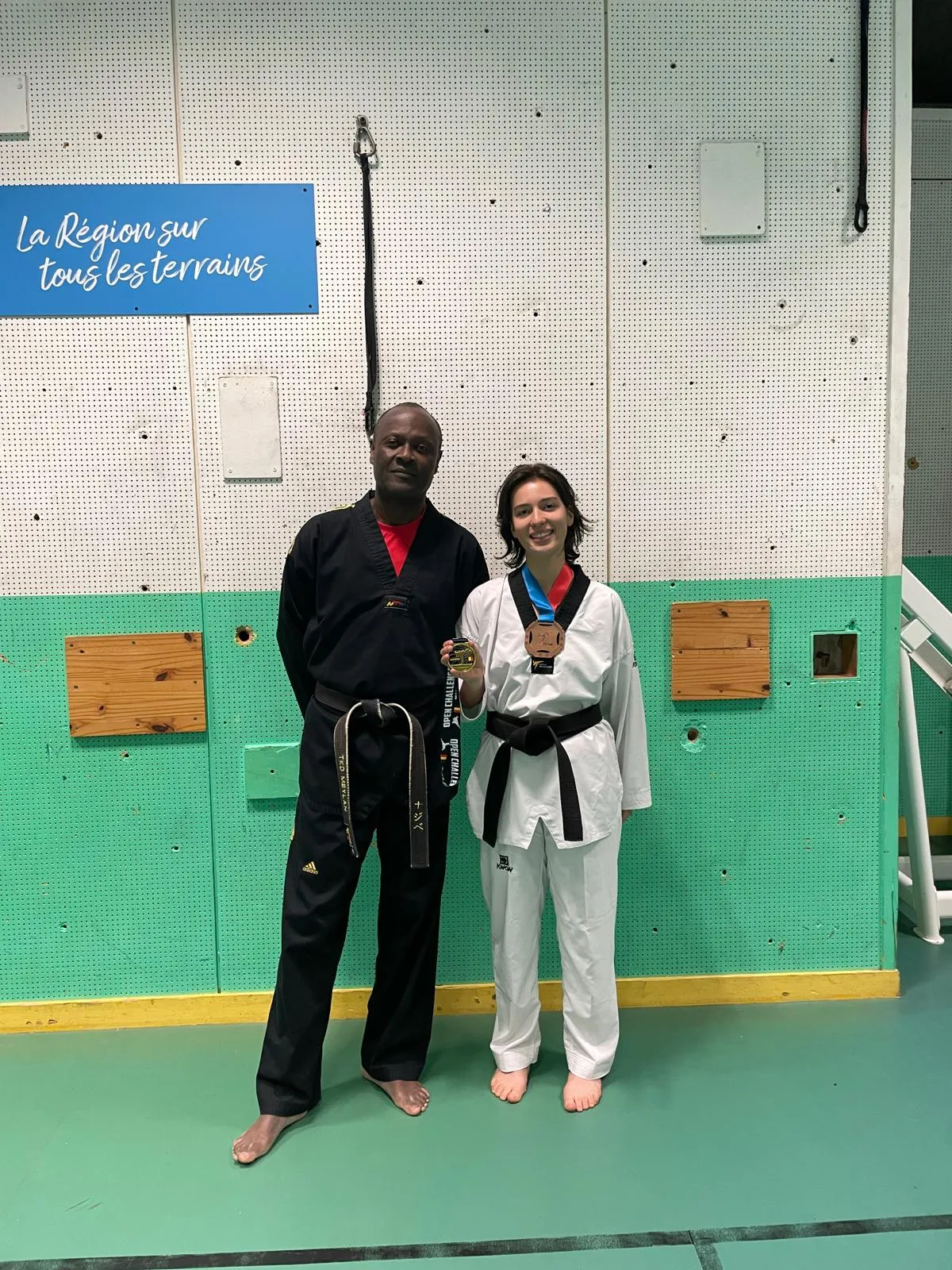 Mila, en dobok et ceinture noire, une médaille autour du cou, aux côtés d'un instructeur du club qui présente une seconde médaille